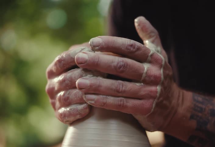 A focused, tattooed artisan with a beard and headphones shapes wet clay on a pottery wheel in slow motion