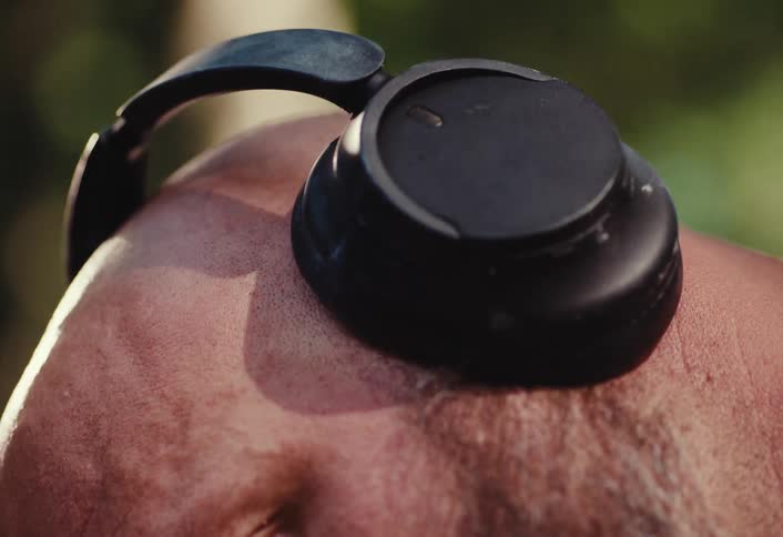 Extreme close-up vertical shot reveals the eye of a bald man with a beard wearing headphones in a natural setting