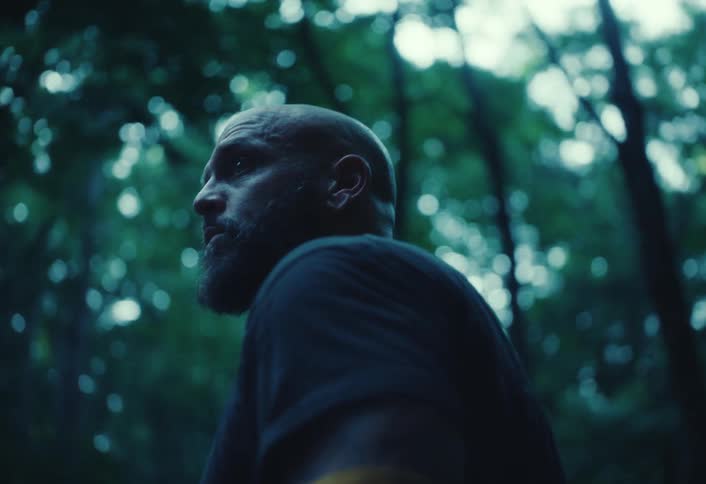 A moody low-angle shot of a serious-looking bald man with a beard standing in a dark forest