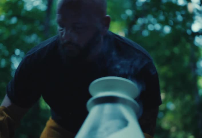 A focused bald craftsman watches over his smoking pottery kiln in a dim outdoor workshop