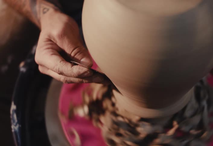 A tattooed artisan carefully shaves excess clay from a spinning vase on a pottery wheel in a slow-motion close-up