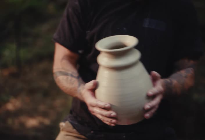 A potter with tattooed arms proudly holds up a freshly made clay pot in an outdoor setting