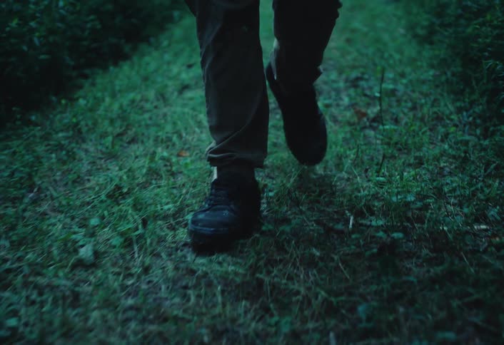 A low-angle shot follows a person's boots as they walk down a grassy path through a dark forest at night