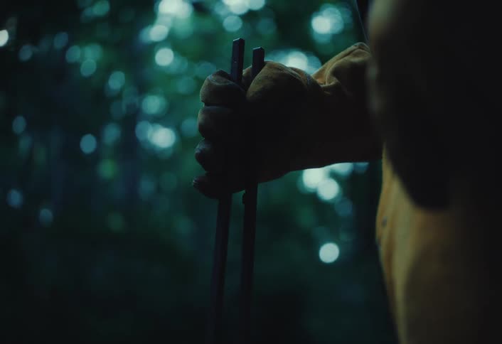 A still, close-up shot of a hand in a yellow work glove firmly holding metal tongs in a dark, mysterious forest
