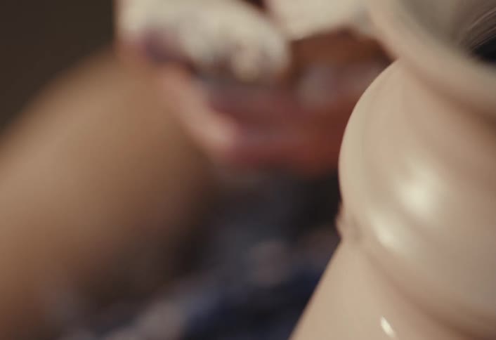A slow-motion close-up shot focuses on a potter's tattooed hands skillfully shaping a wet clay pot on a spinning wheel