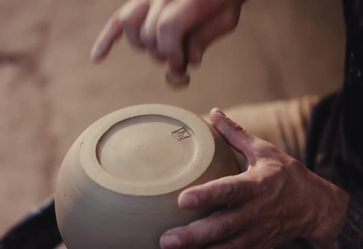 A potter with tattooed arms carefully stamps their signature maker's mark onto the bottom of a handmade clay bowl