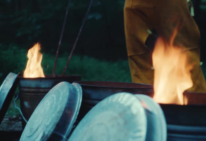 An artisan in protective gear uses tongs to stoke the flames in several metal reduction chambers during an outdoor Raku firing at dusk