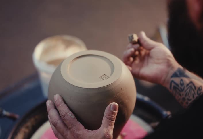 Over-the-shoulder shot of a craftsman's hands carefully stamping a signature mark onto the bottom of a handmade clay pot