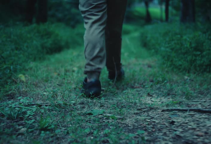 A slow-motion, low-angle shot follows a person's feet as they walk along a grassy path through a dark forest at dusk
