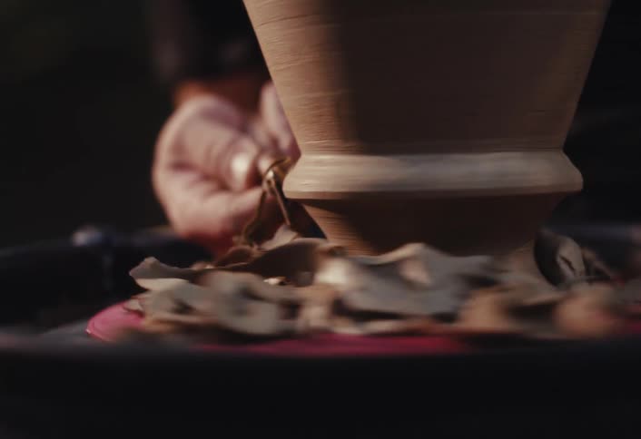 An artisan carefully trims a clay pot on a spinning pottery wheel, with shavings flying off in a detailed slow-motion close-up