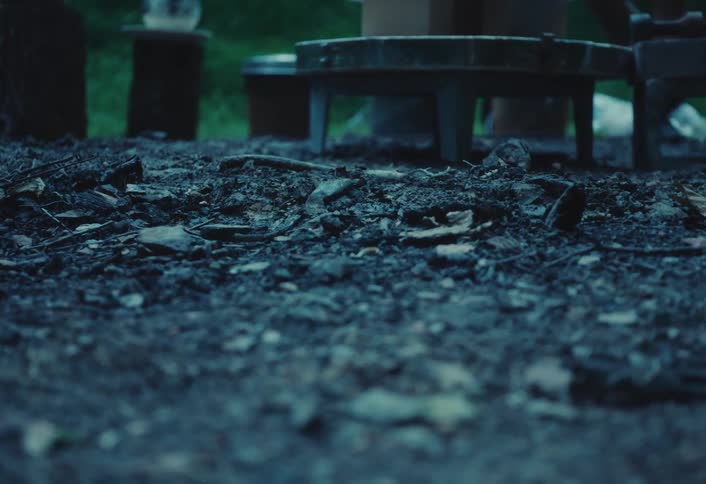 A moody low-angle close-up of a dark, damp forest floor with a campfire pit in the background