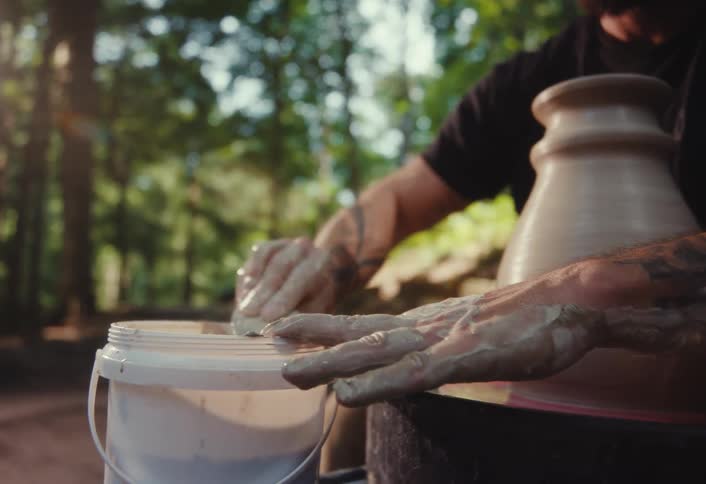 A potter with tattoos cleans his clay-covered hands with a sponge after working on his pottery wheel outdoors in a sunlit forest