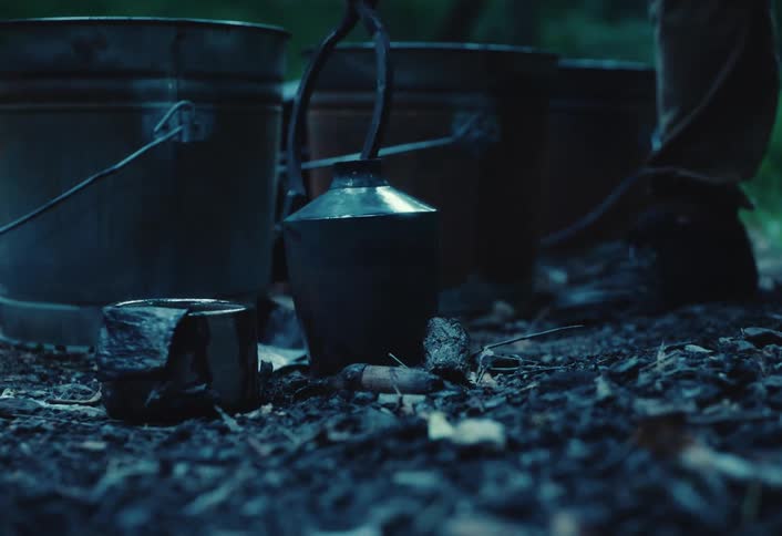 A smoking bee smoker is placed on the dark forest floor next to rustic metal buckets