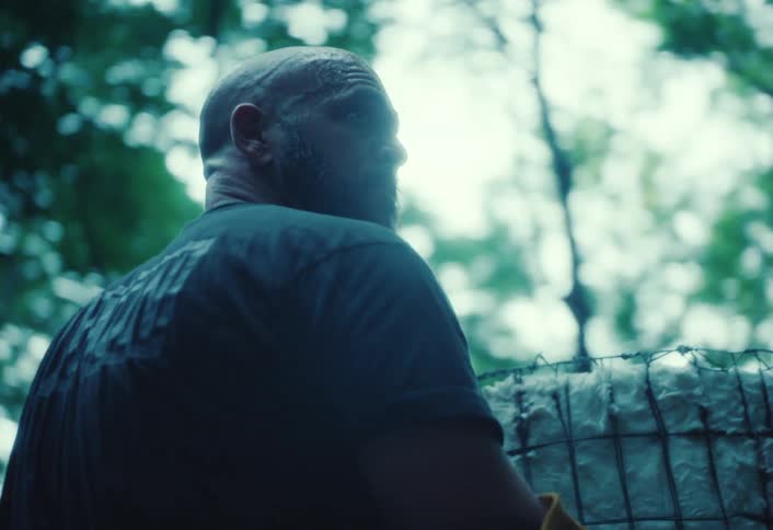 A bearded man in slow motion walks through the woods and carefully unlatches an outdoor kiln
