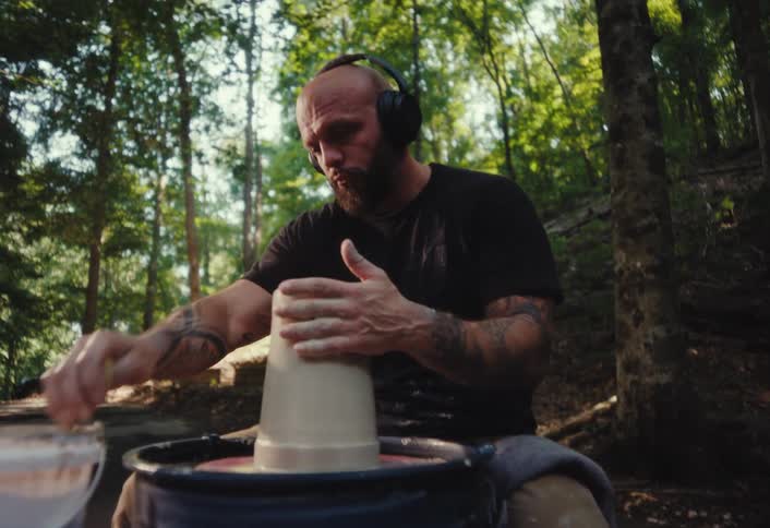 A tattooed man with headphones concentrates on shaping clay on a pottery wheel in a peaceful forest setting