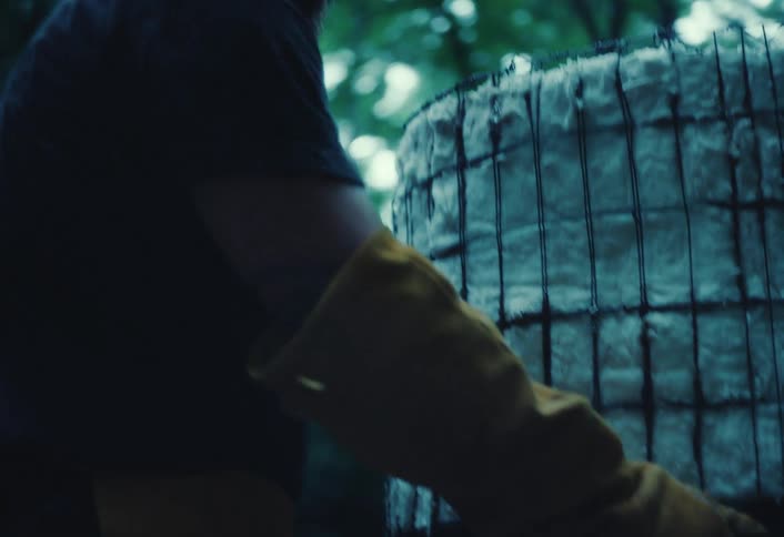 A bearded man wearing protective gloves works on a custom-made outdoor kiln in a moody forest setting