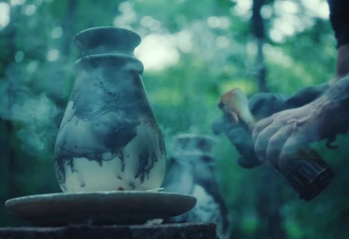 In slow motion, an artisan potter sprays a steaming hot ceramic vase with a finishing glaze outdoors in a forest