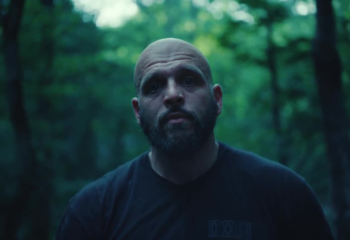 A stoic, bald man with a beard stares intensely at the camera while standing in a dark forest