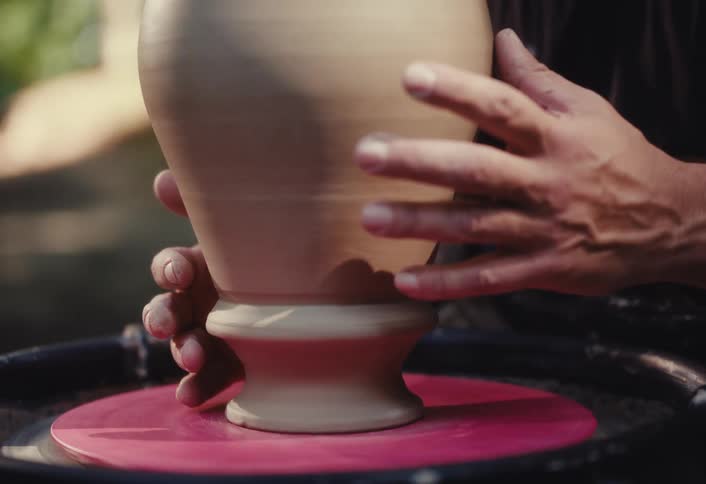 A skilled potter expertly shapes the base of a clay vase on a spinning pottery wheel in a sunlit workshop