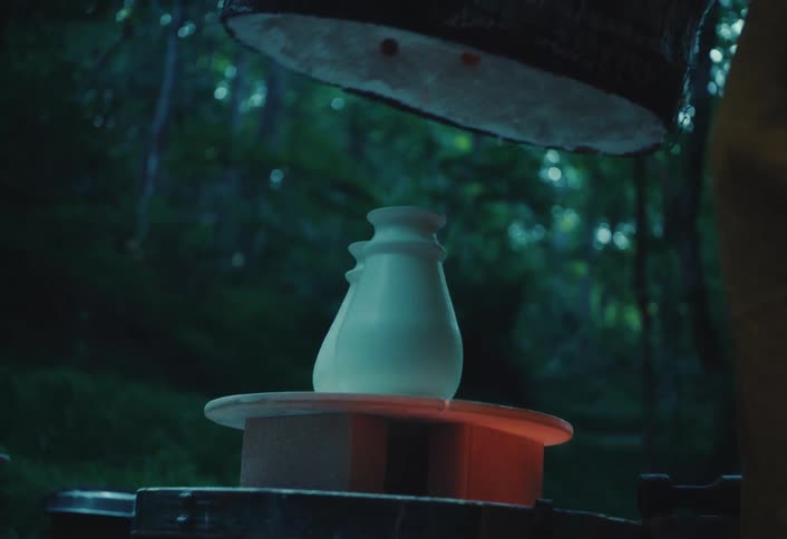 A potter lifts the top of an outdoor kiln to reveal a freshly fired, white ceramic vase glowing from the heat in a forest setting