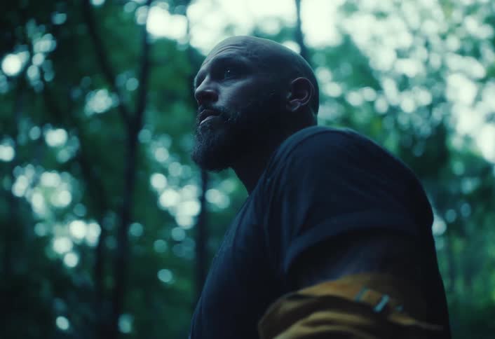 A serious bald man with a beard looks around while standing in a dark, atmospheric forest