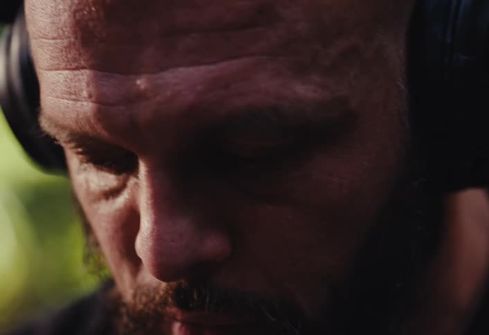 An intense close-up of a bearded man with headphones on, meditating or concentrating while listening to music in a sunlit forest