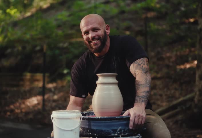 A proud, tattooed potter smiles at the camera next to his freshly made clay vase on a spinning wheel outdoors