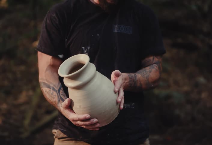 A tattooed craftsman proudly holds his newly created, unfired clay pot in a tranquil forest setting