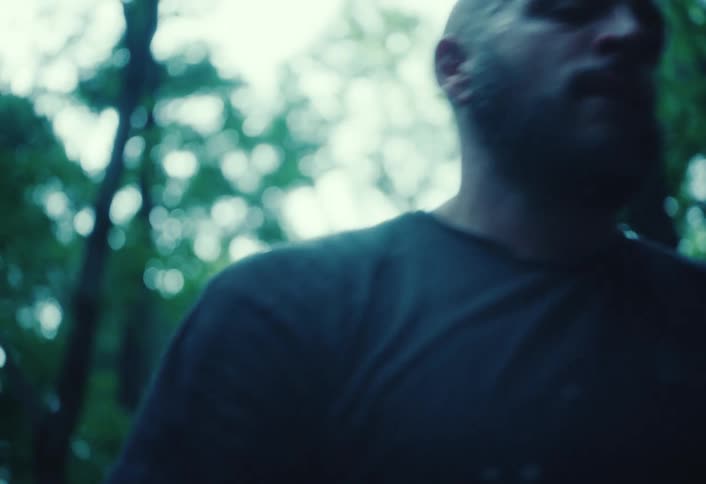 A low-angle shot of an intense, sweaty bald man with a beard standing in a moody forest before walking out of the frame