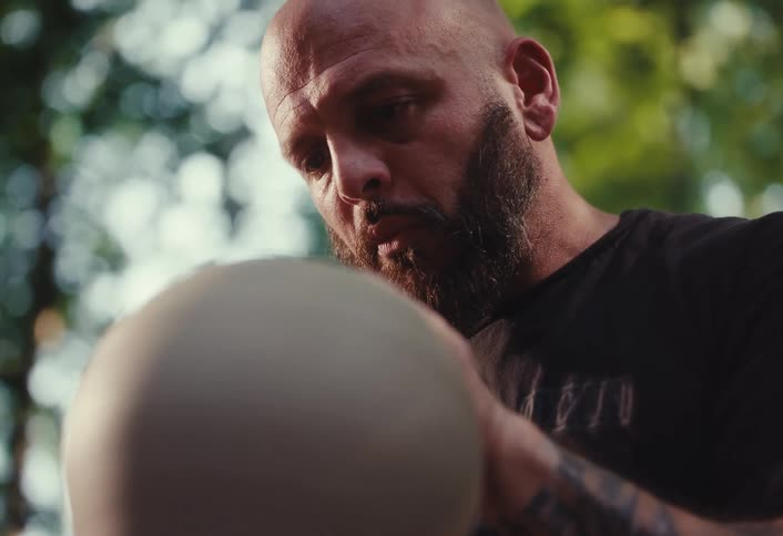 A concentrated, bearded craftsman carefully shapes a piece of clay on a pottery wheel outdoors
