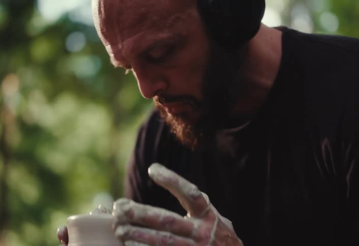 A focused bearded man wearing headphones carefully molds and shapes a clay pot on a pottery wheel in an outdoor setting