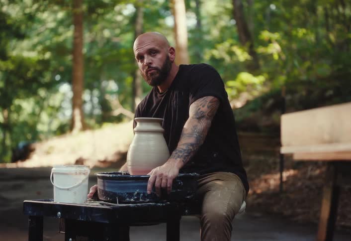 A rugged, tattooed man sits at a potter's wheel shaping a vase, pausing to look seriously into the camera in a slow-motion shot