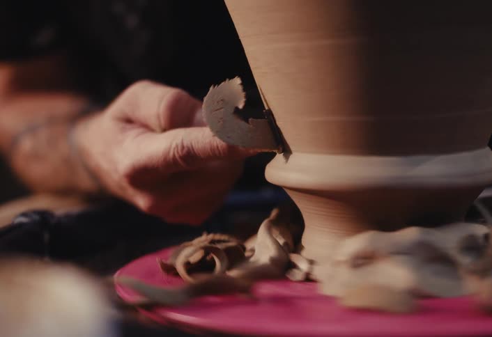 A potter uses a tool to carefully shave and refine a clay pot spinning on a wheel in slow motion