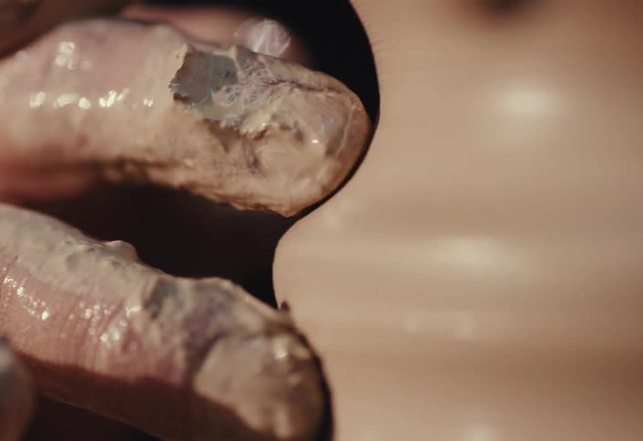 A potter's clay-covered fingers carefully create grooves in a piece of pottery on a spinning wheel in an extreme close-up