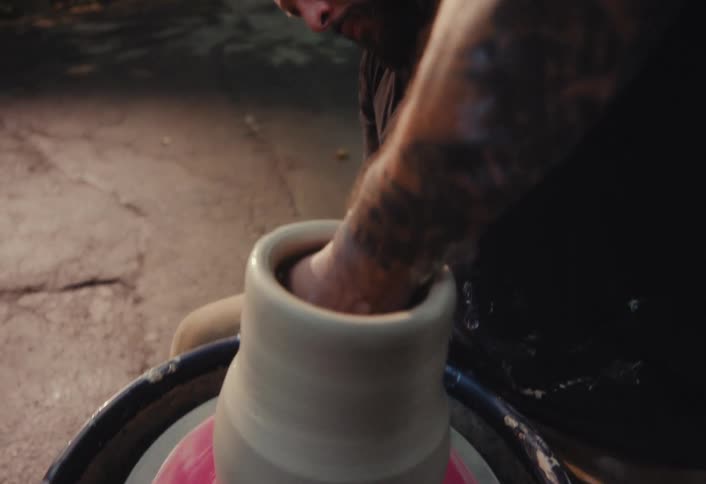 A tattooed man with headphones intently shapes a clay pot on a pottery wheel in an outdoor setting