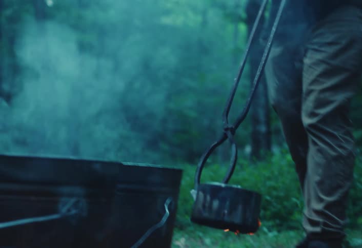 A man wearing a protective glove uses tongs to place a hot crucible from a fire onto the forest floor