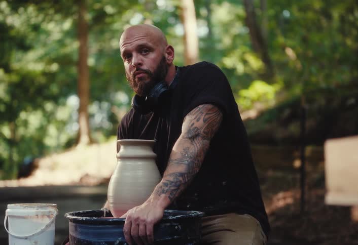 Slow-motion portrait of a tattooed potter looking at the camera while working on his wheel outdoors