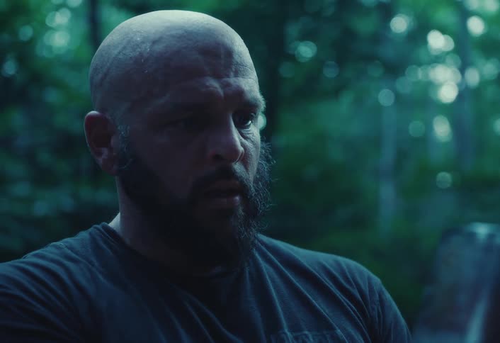 A bearded man looks intently into a steaming hot metal mug while camping alone in a moody, dark forest