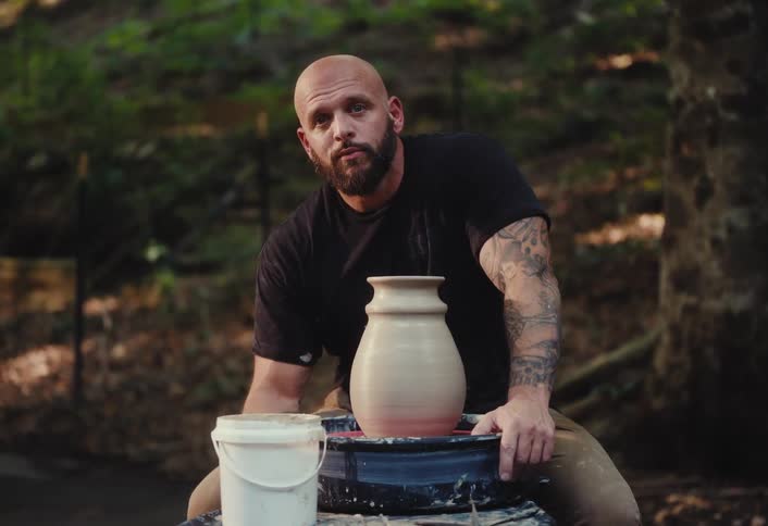 A tattooed potter proudly presents his finished clay vase on a pottery wheel in a serene outdoor setting