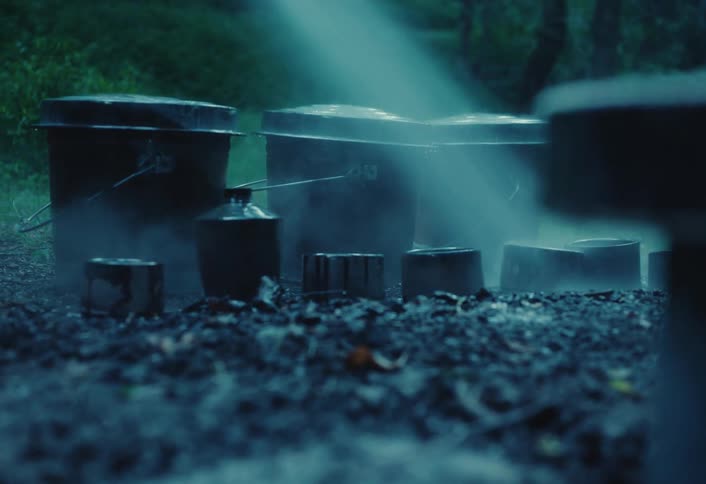 A beam of light illuminates steaming cauldrons on the smoldering ground in a mysterious, dark forest