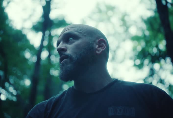 A rugged, bald man with a beard stands in a dense, moody forest, looking around with a serious and intense expression