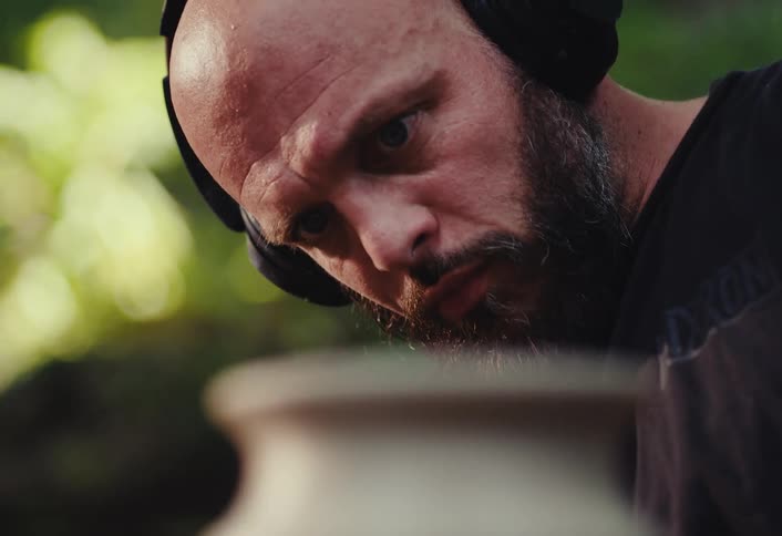 A bald, bearded potter wearing headphones maintains intense focus as he shapes clay on a potter's wheel in his outdoor studio
