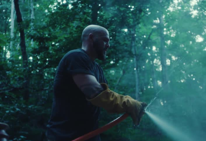 A rugged man with work gloves sprays water from a hose in a moody, green forest