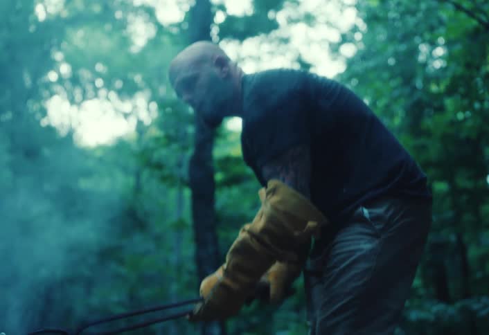 A craftsman in a smoky forest carefully uses tongs to place a red-hot crucible onto the ground