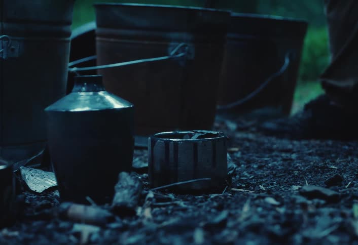A person in a dark forest uses tongs to carefully place a small pot with glowing embers on the ground next to old metal buckets
