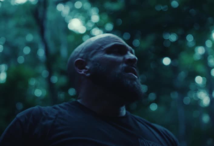 A rugged, bald man working in a dark forest stops to wipe sweat from his brow in slow motion