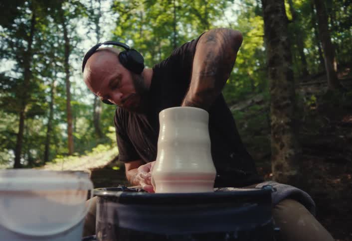 A tattooed potter wearing headphones intently works on a piece of clay on a pottery wheel outdoors in the woods