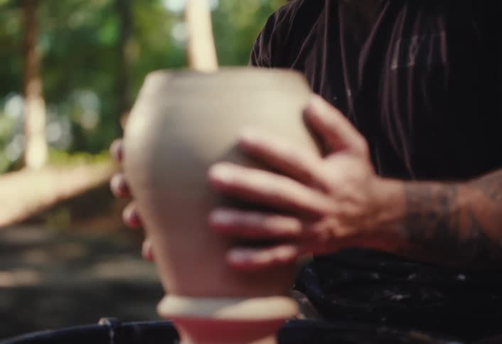 A potter with tattooed arms carefully removes a clay vase from the spinning wheel and places it back to continue shaping in slow motion