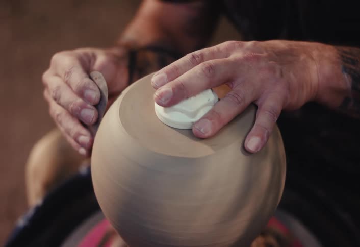 A skilled artisan with tattoos carefully shapes a clay vase on a spinning pottery wheel in his workshop