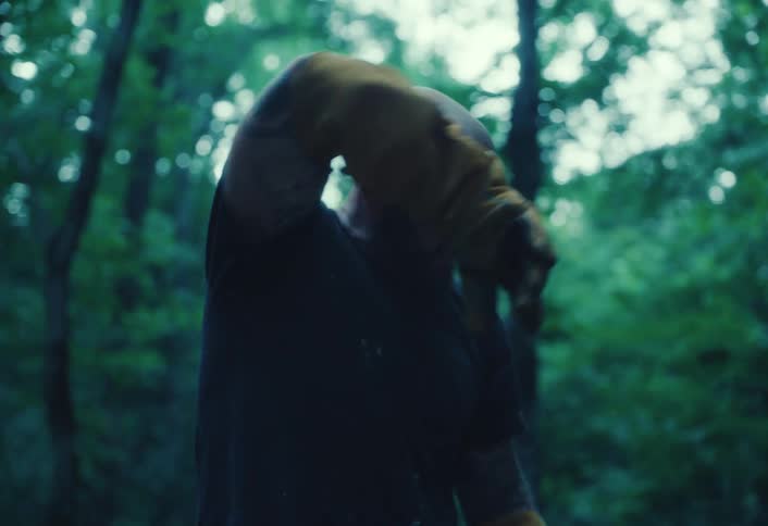 A muscular, tattooed man in a forest wipes the sweat from his brow with his gloved hand in slow motion